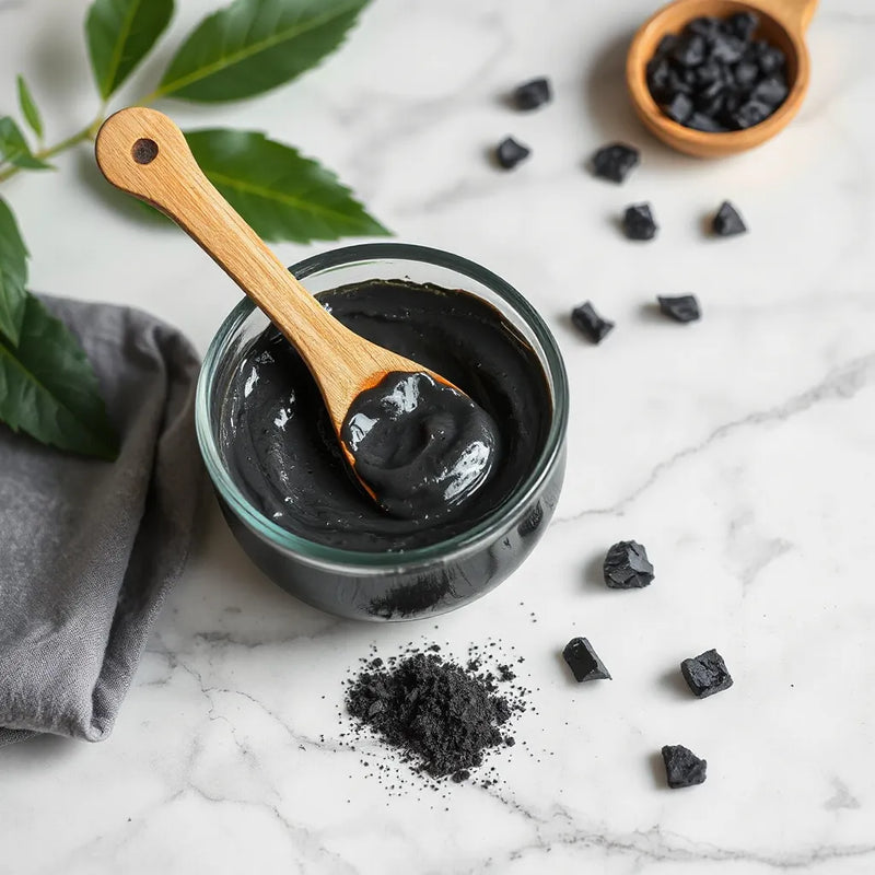 Charcoal mask in a bowl with a wooden spatula and brush on a neutral skincare surface